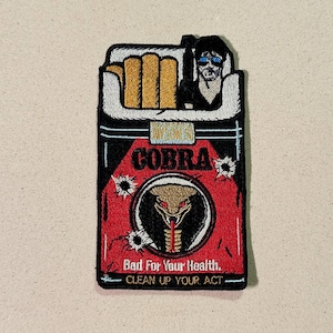 Peut inclure: Écusson brodé représentant un paquet de cigarettes avec le mot "COBRA" en rouge. L'écusson comprend un emblème de serpent et le texte "Bad For Your Health. CLEAN UP YOUR ACT."