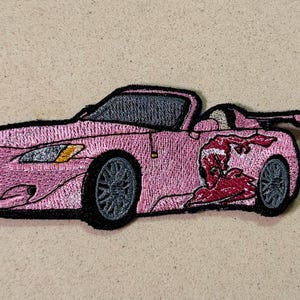May include: Pink embroidered patch of a convertible sports car with black trim and grey wheels. The car has a red graphic on the side. A ruler shows the patch is approximately 5 cm in length.