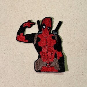 以下が含まれることがあります： ダイナミックなポーズのデッドプールのキャラクターが描かれた刺繍パッチ。パッチは、デッドプールの象徴的な赤と黒のスーツと、背中に2本の刀剣を表示しています。デザインには、詳細なステッチとベルトバックルが含まれています。