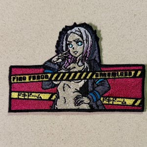 Può includere: Toppa ricamata raffigurante un personaggio femminile di anime con occhi blu e capelli rosa, che indossa una giacca con cappuccio. La toppa ha uno sfondo rosso con strisce di pericolo gialle e nere e il testo "FIRE FORCE" e caratteri giapponesi.