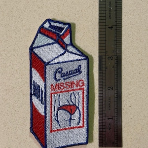 以下が含まれることがあります： 牛乳パックの形をした刺繍パッチ。パッチは主にシルバーで、赤と青のアクセントがあります。「Casual MISSING」というテキストは、赤い下着を身に着けた人の後ろ姿のイラストの上に表示されています。