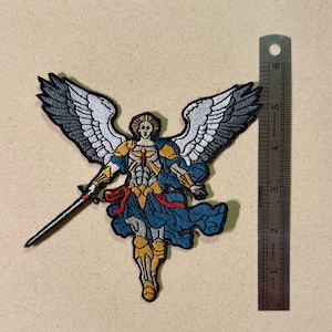 May include: Embroidered patch featuring an angel with outstretched wings, holding a sword. The angel is depicted in blue, gold, and red, with white and gray wings. A metal ruler is next to the patch, showing inches.