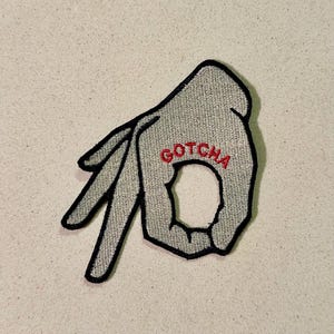 Puede incluir: Parche bordado con la forma de una mano haciendo el gesto de 'ok'. El parche es gris con contorno negro y la palabra 'GOTCHA' en rojo. Adecuado para ropa o accesorios.