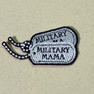 May include: Embroidered patch in the shape of a military dog tag, featuring the words "MILITARY" and "MILITARY MAMA" in a silver color with a black border. The patch has a chain-like detail.