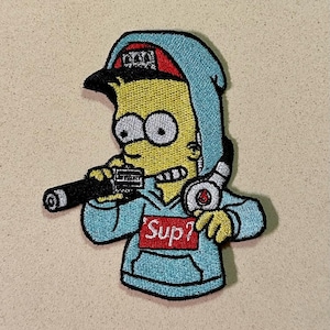 Pode incluir: Patch bordado com um personagem de desenho animado usando um moletom com capuz e boné azuis, segurando um objeto preto. O personagem é amarelo com olhos grandes e uma expressão travessa. O moletom tem a palavra "Sup?" nele.