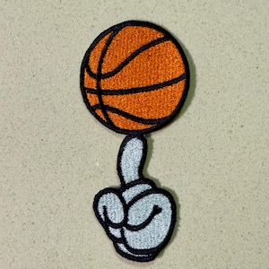 Könnte beinhalten: Gestickter Aufnäher mit einem Basketball, der auf einer Cartoon-Hand balanciert. Der Basketball ist orange mit schwarzen Linien, und die Hand ist grau mit schwarzem Umriss. Ein lustiges, sportliches Design.