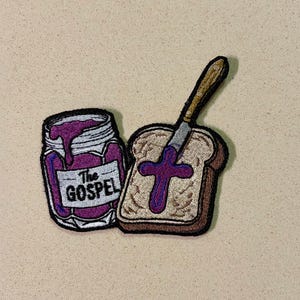 Puede incluir: Parche bordado con un tarro de mermelada morada con el texto "The Gospel" y una rebanada de pan con una cruz de mermelada y un cuchillo. El parche tiene un borde blanco.