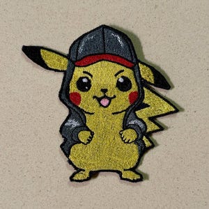 May include: Embroidered patch featuring a yellow Pikachu character wearing a black hoodie and a red-billed cap. The patch has black eyes, red cheeks, and a small pink tongue. The design is outlined in black.