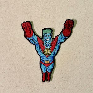 May include: Embroidered patch of a superhero with blue skin, green hair, and red gloves and boots. The superhero has a red chest and waist band with a gold globe emblem. The patch is on a beige background.