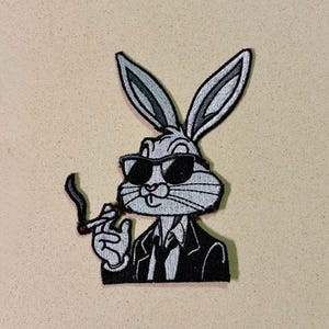 May include: Embroidered patch featuring a cartoon rabbit wearing sunglasses and a black suit, holding a cigarette. The rabbit is gray and white with black outlines. The patch is on a beige background.
