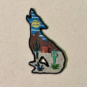 Pode incluir: Patch bordado em forma de lobo uivando, com uma paisagem desértica. O lobo é delineado em preto, com um céu azul e uma lua amarela. A cena do deserto inclui cactos e um edifício.