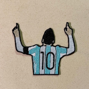May include: Embroidered patch featuring a soccer player in a light blue and white striped jersey with the number 10 on the back. The player is depicted with arms raised, index fingers pointing upwards, and a black silhouette for the head.