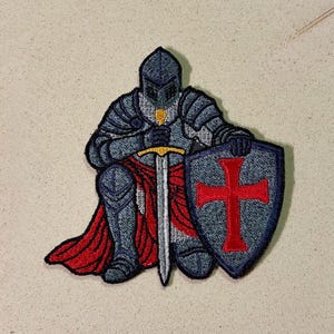 May include: Embroidered patch featuring a kneeling knight in full armor. The knight is holding a sword and shield with a red cross. The knight's armor is blue and gray, with a red cape.