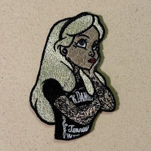 May include: Embroidered patch featuring a stylized illustration of a woman with light blonde hair, blue eyes, and red lips. She is wearing a black t-shirt with the text "Jack Daniels Tennessee Whiskey" and has tattoos on her arms.