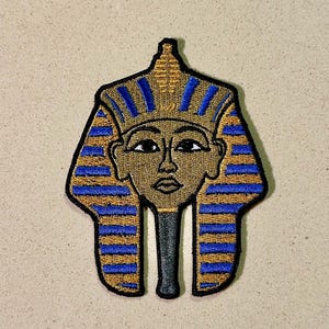 Könnte beinhalten: Gestickter Aufnäher mit dem stilisierten Gesicht eines ägyptischen Pharaos. Der Aufnäher hat ein Gold- und Blau-Farbschema mit schwarzer Umrandung. Das Gesicht des Pharaos ist braun mit schwarzen Augen und Lippen. Ein einzigartiges Accessoire.