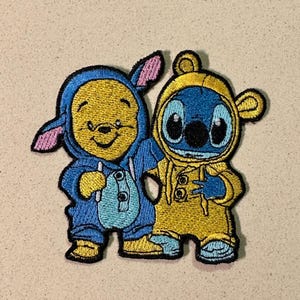 May include: Embroidered patch featuring two cartoon characters in costumes. One is yellow and blue, resembling a bear, and the other is blue and yellow, resembling a koala. The patch has a black border and is likely for clothing or accessories.