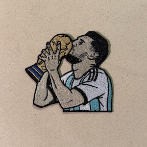 May include: Embroidered patch featuring a soccer player kissing a gold trophy. The player is wearing a white and light blue jersey. The patch has a black border and is on a tan background.