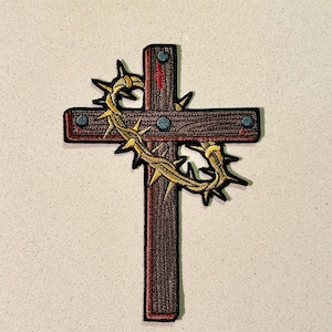 May include: Embroidered patch depicting a brown cross with a gold crown of thorns. The cross has a red accent and blue studs. The patch has a black border, suitable for application on clothing or accessories.