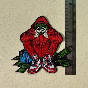 May include: Embroidered patch featuring a figure in a red hooded jumpsuit, hands covering the face, holding green money. The figure is seated with red and white sneakers. A blue bag and green money are also present. The patch measures approximately 3 inches.
