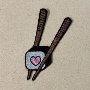 Op de afbeelding: Geborduurde patch met een sushirol met een roze hart in het midden, geflankeerd door twee bruine eetstokjes. De sushirol is lichtblauw en zwart. De patch staat op een beige achtergrond.