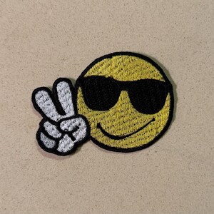 May include: Embroidered patch featuring a yellow smiley face wearing black sunglasses and giving a peace sign with its left hand. The patch has a black border and is on a tan background.