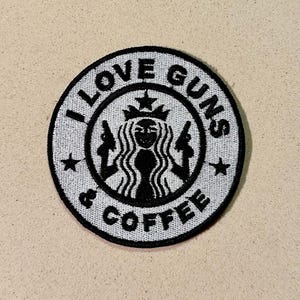 May include: A circular patch with the text "I LOVE GUNS & COFFEE" in a white, stylized font. The design features a black silhouette of a woman with guns in each hand, surrounded by a white border and stars.