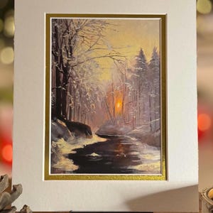 May include: A framed landscape painting of a snowy forest scene. The artwork depicts a river winding through snow-covered trees, with a warm sunset in the background. The painting is set within a white mat and a gold frame.