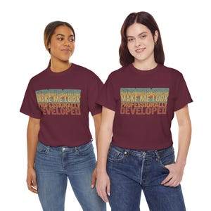 May include: A maroon short-sleeved t-shirt featuring a retro-style graphic print with the text DOES THIS SHIRT MAKE ME LOOK PROFESSIONALLY DEVELOPED in stacked lines of blue, yellow, and orange against a dark background.