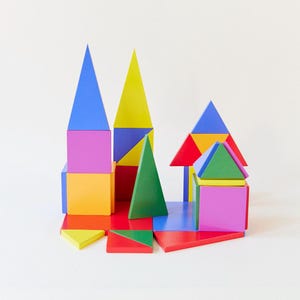 Puede incluir: Un colorido juego de bloques de madera con varias formas geométricas, incluyendo triángulos y cubos. Los bloques están pintados en colores vibrantes como azul, amarillo, verde, rojo y morado, dispuestos para parecerse a una construcción.