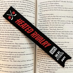 May include: Black bookmark with the text "HEATED RIVALRY" in red, featuring hockey sticks and a puck graphic. The bottom has a VHS tape design with "Hi-Fi STEREO 2471".