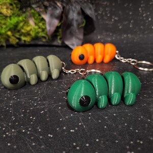 Puede incluir: Tres llaveros de oruga articulados en verde oliva, naranja brillante y verde bosque. Cada figura de insecto segmentada presenta una cabeza redondeada y lisa con un ojo circular negro prominente y una anilla de metal en la parte posterior.