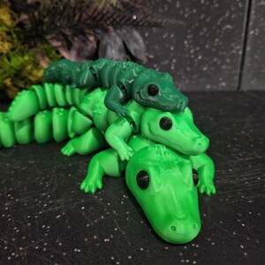 Puede incluir: Una pila de tres figuras de caimán articuladas en varios tonos de verde. El caimán superior es verde oscuro, el del medio es verde medio y el inferior es verde lima brillante. Cada figura presenta ojos negros y un cuerpo segmentado.