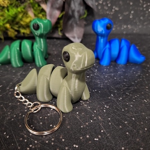 Puede incluir: Tres figuras de dinosaurios articuladas en verde, azul y verde oliva. Cada criatura presenta un cuerpo liso y segmentado con una cabeza redondeada y grandes ojos negros. El dinosaurio verde oliva está unido a un llavero de metal plateado.