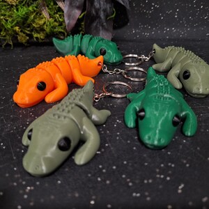 Puede incluir: Una colección de cinco llaveros de caimán articulados en naranja, verde brillante y verde oliva apagado. Cada caimán presenta un cuerpo segmentado, una espalda texturizada y ojos negros. Los llaveros están unidos a anillas metálicas plateadas.