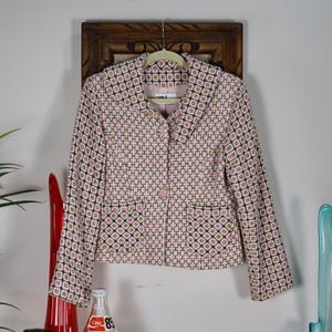 May include: A pink, black, and tan patterned blazer with a geometric design. The blazer has long sleeves, two front pockets, and a single-button closure. The blazer is hanging on a wooden hanger.