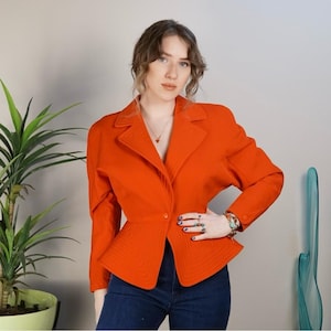 May include: A bright orange blazer with a fitted waist and a slightly flared hem. The blazer has a single button closure and a pointed collar.