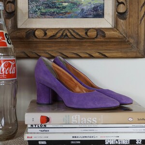 May include: A pair of purple suede heels with a block heel. The shoes are displayed on a stack of books, with a vintage Coca-Cola bottle and a framed painting in the background.