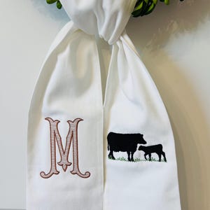 May include: A white fabric sash featuring an embroidered brown letter M on the left side and a black cow and calf silhouette standing on green grass on the right side. The fabric is knotted at the top and hangs down in two separate panels.
