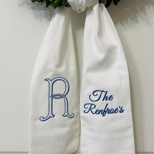 May include: White fabric sash with blue embroidered text and a green wreath. The sash features the letter "R" and the text "The Renfroe's". The sash is tied to a green wreath, creating a decorative element.