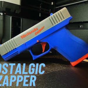 May include: A blue, gray, and orange Nintendo Zapper toy gun. The words "Nintendo Zapper" are printed in orange on the gray top. The text "NOSTALGIC ZAPPER" is printed in blue at the bottom. The toy gun is angled to the left.