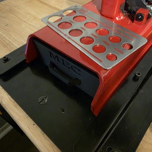 May include: A red and black MEC reloading press with a silver plate featuring numbered holes. The press sits on a wooden surface. The MEC logo is visible on the front of the press.