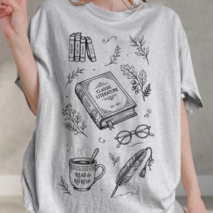 May include: Light gray t-shirt featuring a black and white illustration of books, reading glasses, a coffee cup with the text "Read & Refresh", and a feather quill. The shirt design includes the text "Classic Literature" on a book.