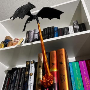 May include: A black dragon figurine with outstretched wings positioned above a long, vertical, translucent orange flame effect that extends downward to a textured orange base, creating a dynamic display of a dragon breathing fire.