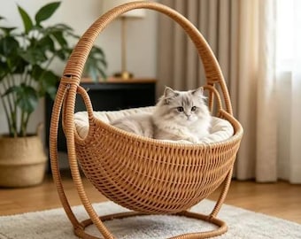 May include: A light brown woven rattan pet bed with a curved handle and a circular base. The bed features a soft, cream-colored cushion inside, currently occupied by a fluffy, long-haired cat with grey and white fur sitting comfortably.