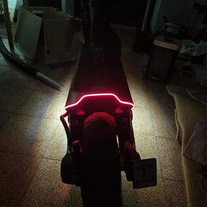 Puede incluir: Un patinete eléctrico negro visto desde atrás, con una llamativa tira de luz LED roja en la base. El patinete tiene un neumático grueso, un mástil vertical y un pequeño soporte rectangular para matrícula con el texto etsy impreso.