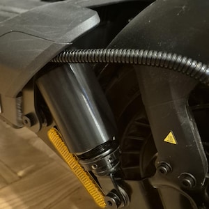 Op de afbeelding: Close-up van het veersysteem van een zwarte elektrische scooter. De afbeelding toont de schokdemper, een gele reflector en een waarschuwingssymbool. Een zwarte kabel loopt langs het frame.