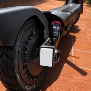 Puede incluir: Un patinete eléctrico negro con una rueda trasera que tiene un neumático 60-7.5, una llanta negra y un reflector rectangular blanco montado en el lateral. Un amortiguador de suspensión rojo y negro es visible sobre la rueda, unido al chasis.