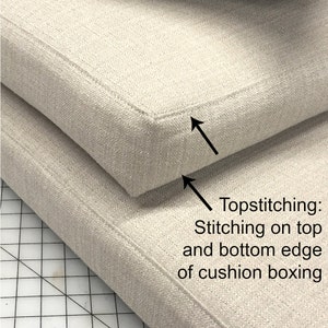 May include: Close-up of two beige fabric cushions with topstitching on the top and bottom edges. The topstitching is a decorative stitch that runs along the edge of the cushion.