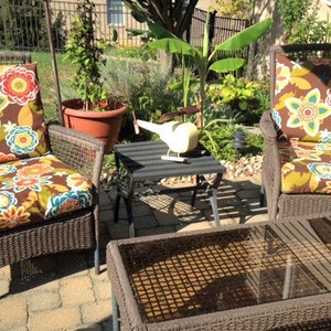 May include: Two wicker chairs with floral patterned cushions and a glass-top table on a patio. The chairs are brown and the cushions have a brown, yellow, orange, and blue floral pattern. The table is brown with a glass top. There is a small toy helicopter on a black table in the background.