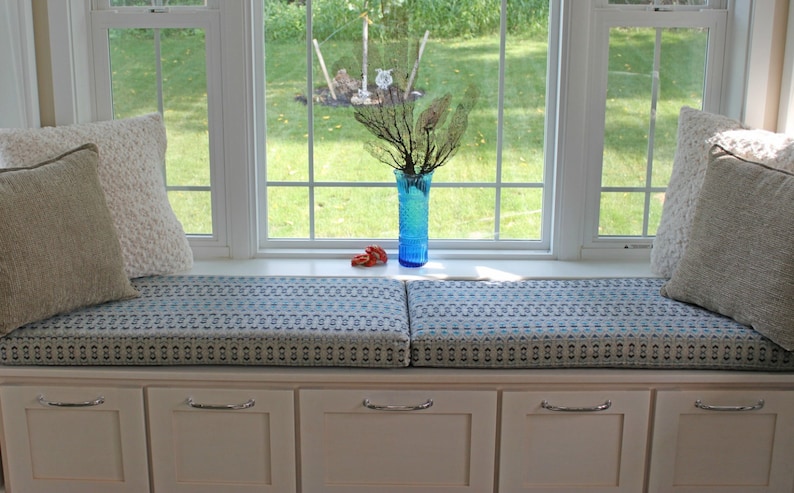 May include: A white window seat with a blue and white patterned cushion. The seat has two cushions and is flanked by white cabinets with silver handles. A blue glass vase with a dried branch sits on the window sill.